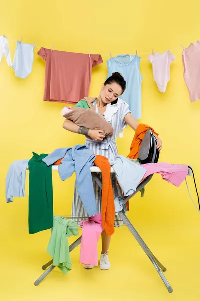 Busy woman talking on cellphone while holding baby doll and iron near pile of laundry on yellow — Stock Photo