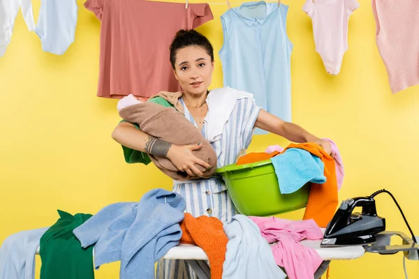 Busy woman looking at camera while holding baby doll and laundry bowl on yellow — Stock Photo