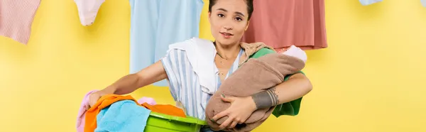 Hausfrau blickt in die Kamera, während sie Babypuppe und Wäsche in der Nähe von Kleidung hält, die auf gelbem Hintergrund hängt, Banner — Stockfoto