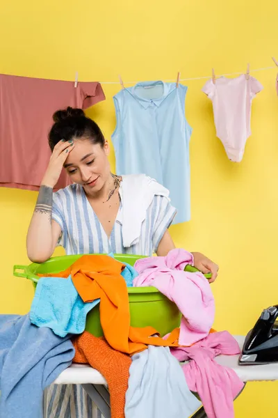 Exhausto ama de casa tocando la cabeza mientras está de pie cerca de lavandería en tabla de planchar en amarillo - foto de stock