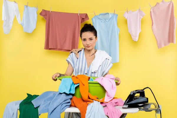 Молода домогосподарка дивиться на камеру біля пральні та прасувальної дошки на жовтому — стокове фото
