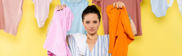 Housewife looking at camera while holding laundry near clothing hanging on yellow background, banner — Stock Photo