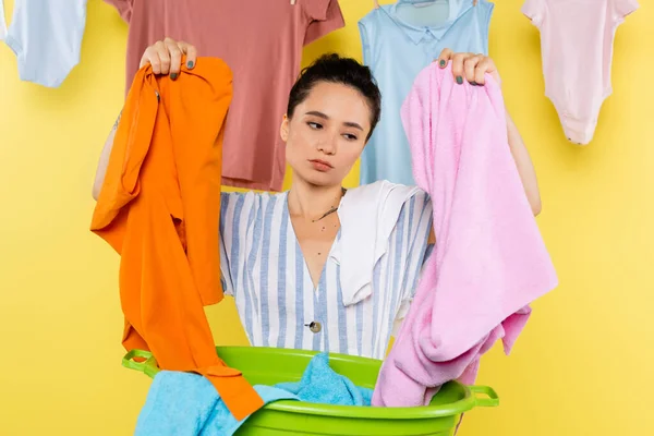 Exhausted housewife holding clothes near laundry bowl on yellow background — Stock Photo