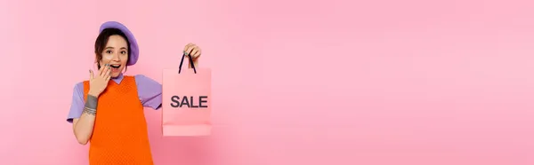 Astonished woman with sale shopping bag covering open mouth with hand isolated on pink, banner — Stock Photo