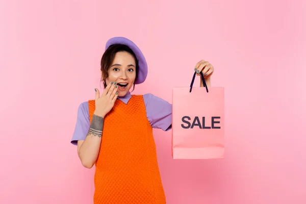 Femme étonnée couvrant bouche ouverte avec la main tout en montrant sac à provisions de vente isolé sur rose — Photo de stock