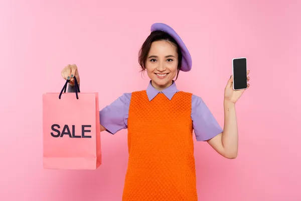 Mujer alegre en vestido anaranjado sin mangas que muestra el teléfono inteligente y la venta bolsa aislada en rosa - foto de stock
