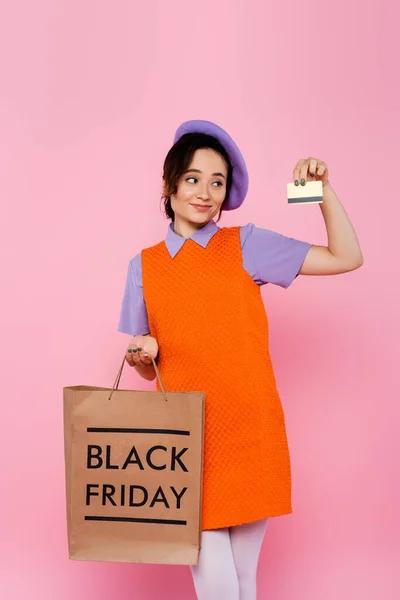 Menina satisfeito em boina roxa segurando cartão de crédito e saco de compras com letras sexta-feira preto isolado em rosa — Fotografia de Stock