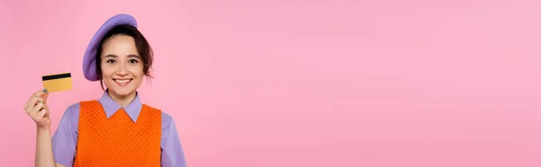 Felice donna in berretto viola sorridente alla macchina fotografica mentre mostra la carta di credito isolata su rosa, banner — Foto stock