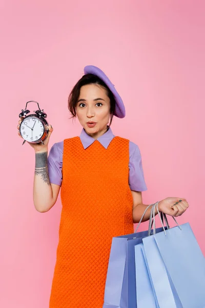 Amazed woman with shopping bags showing vintage alarm clock isolated on pink — Stock Photo