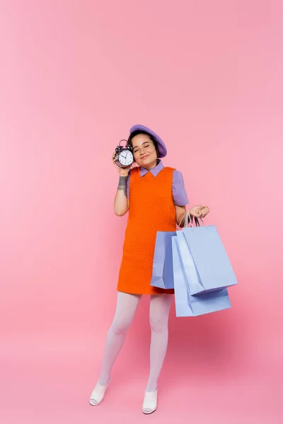 Mujer complacida con los ojos cerrados sosteniendo reloj despertador vintage y coloridas bolsas de compras en rosa - foto de stock