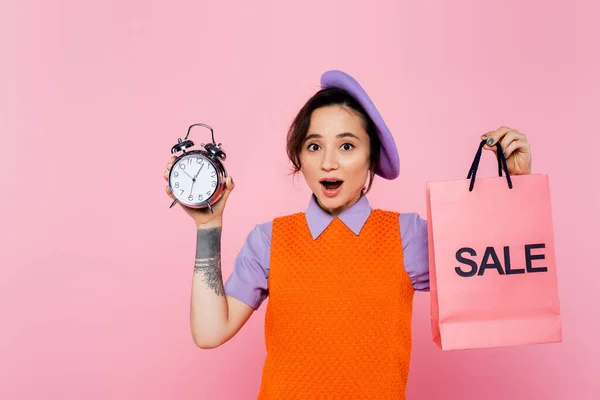 Excited woman in bright clothes holding vintage alarm clock and sale shopping bag isolated on pink — Stock Photo