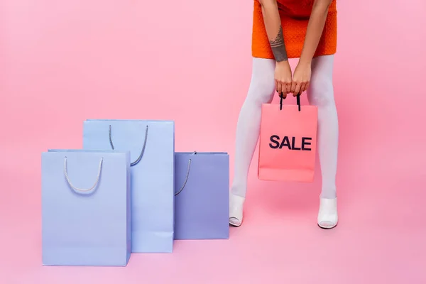 Partial view of tattooed woman holding shopping bag with sale lettering near purchases on pink — Stock Photo