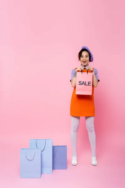 Erstaunte Frau zeigt Einkaufstasche mit Verkaufsaufdruck bei Einkäufen auf rosa — Stockfoto
