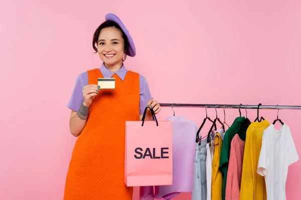 Mujer con estilo con tarjeta de crédito y bolsa de compras con letras venta sonriendo cerca de prendas aisladas en rosa - foto de stock