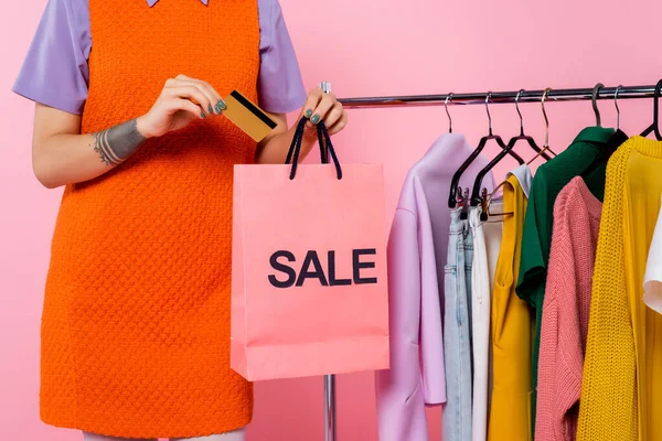 Vista ritagliata di donna tatuata con carta di credito e vendita shopping bag vicino a rack con capi isolati su rosa — Foto stock