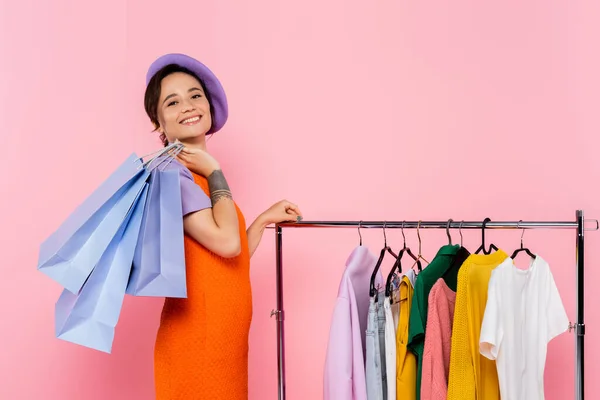 Mujer alegre y de moda con bolsas de compras mirando a la cámara cerca de rack con ropa aislada en rosa - foto de stock