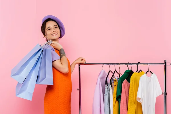 Donna gioiosa e alla moda con borse della spesa guardando macchina fotografica vicino rack con vestiti isolati su rosa — Foto stock