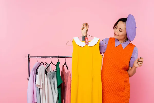 Mulher sorrindo segurando vestido amarelo sem mangas com etiqueta de venda perto rack com roupas isoladas em rosa — Fotografia de Stock