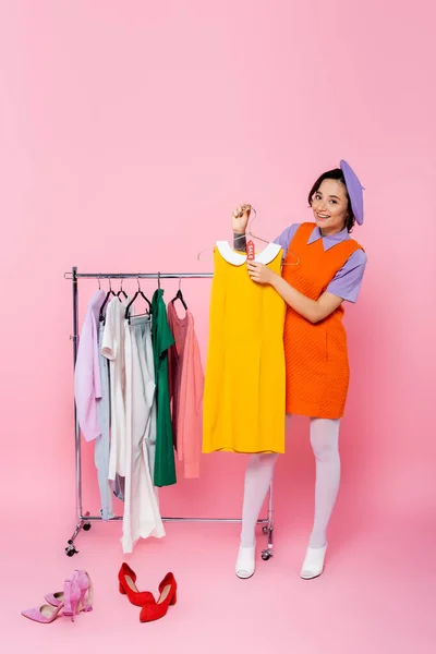 Mulher feliz segurando vestido sem mangas amarelo com venda tag perto de calçado e rack com roupas em rosa — Fotografia de Stock