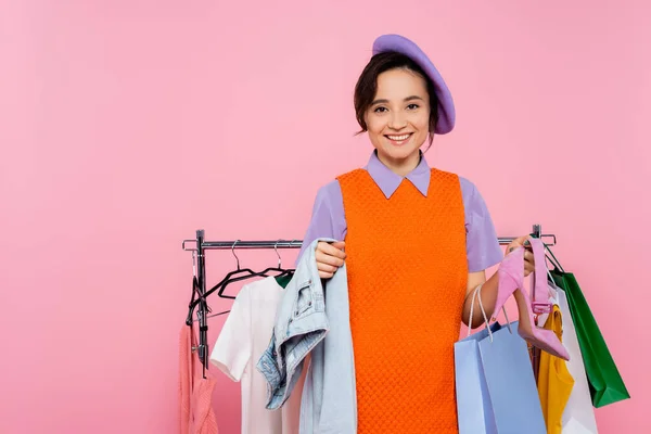 Trendy woman with clothes and shopping bag smiling at camera isolated on pink — Stock Photo