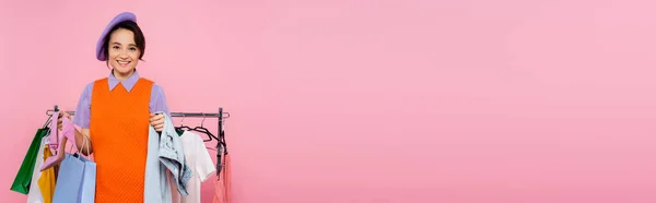 Mujer alegre con bolsa de compras y surtido de ropa mirando a la cámara aislada en rosa, pancarta - foto de stock