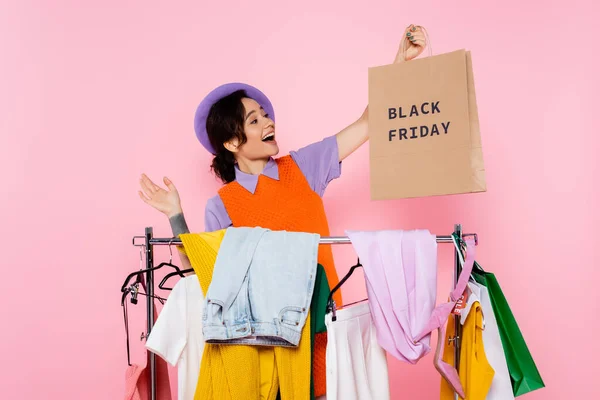 Mujer asombrada sosteniendo bolsa de compras con letras de viernes negro cerca de rack con ropa aislada en rosa - foto de stock