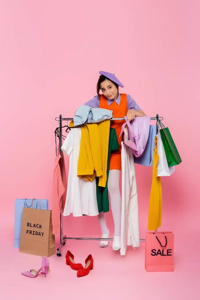 Woman in purple beret looking at camera near rack with garments and shopping bags on black friday sale on pink — Stock Photo