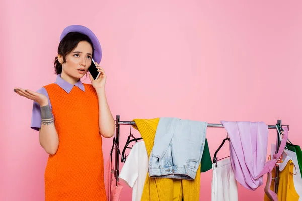 Verärgerte Frau spricht auf Smartphone in der Nähe von Regal mit Kleidung isoliert auf rosa — Stockfoto