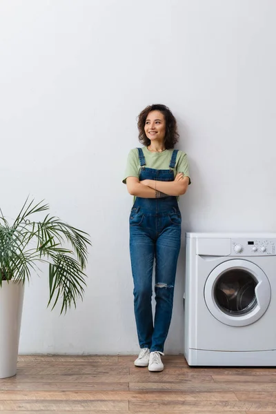 Sorridente dona de casa de pé com os braços cruzados perto de máquina de lavar roupa e planta em casa — Fotografia de Stock