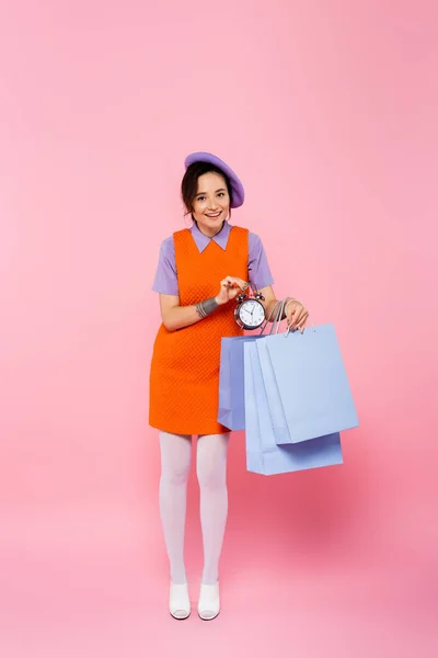 Mujer feliz en vestido anaranjado sin mangas sosteniendo bolsas de compras y reloj despertador vintage en rosa - foto de stock