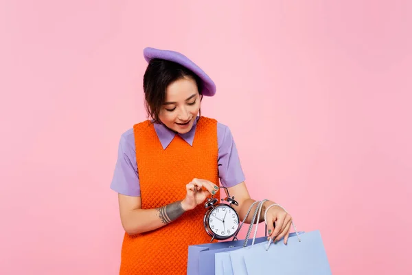 Trendy woman in orange sleeveless dress putting vintage alarm clock into shopping bag isolated on pink — Stock Photo