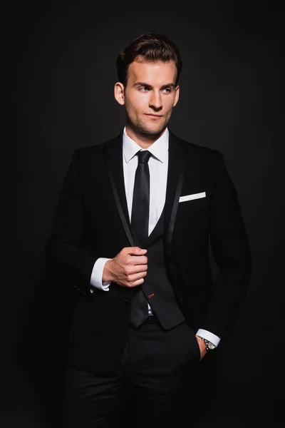 Young man in stylish suit looking away while standing with hand in pocket isolated on black — Stock Photo