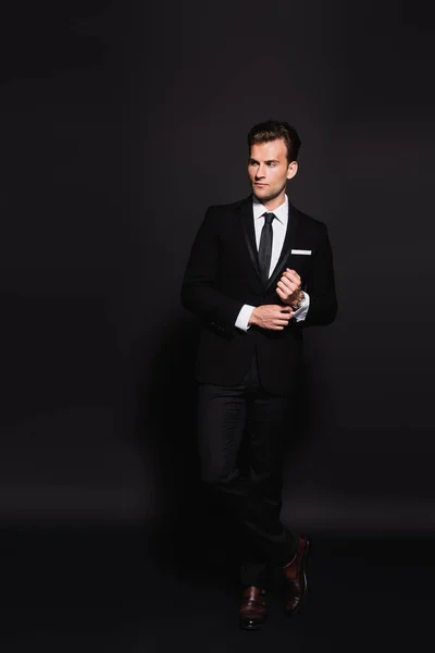 Full length view of young man in elegant suit touching sleeve of white shirt on black — Stock Photo