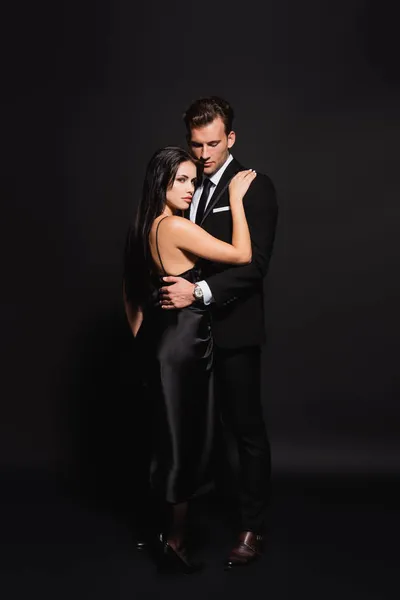 Visão de comprimento total do homem de terno abraçando mulher elegante em vestido de cetim em preto — Fotografia de Stock