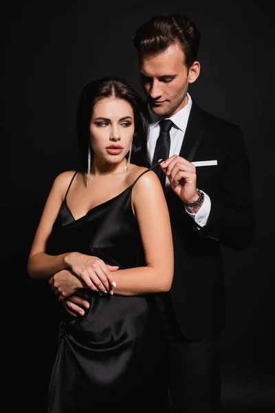 Man in suit touching strap of silk dress on sexy woman while seducing her on black — Stock Photo