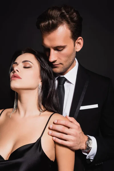 Elegante hombre tocando hombro de mujer seductora de pie con los ojos cerrados aislados en negro - foto de stock