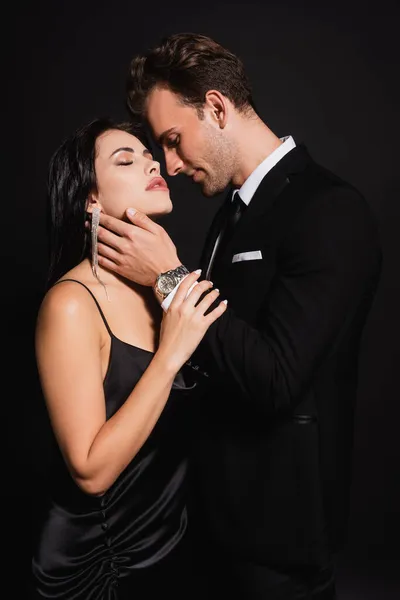 Leidenschaftliche Frau, die mit geschlossenen Augen in der Nähe eines eleganten Mannes steht, der ihren Hals isoliert auf schwarz berührt — Stockfoto