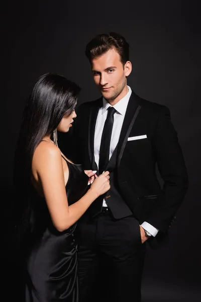 Sensual brunette woman unbuttoning vest of elegant man standing with hand in pocket on black — Stock Photo