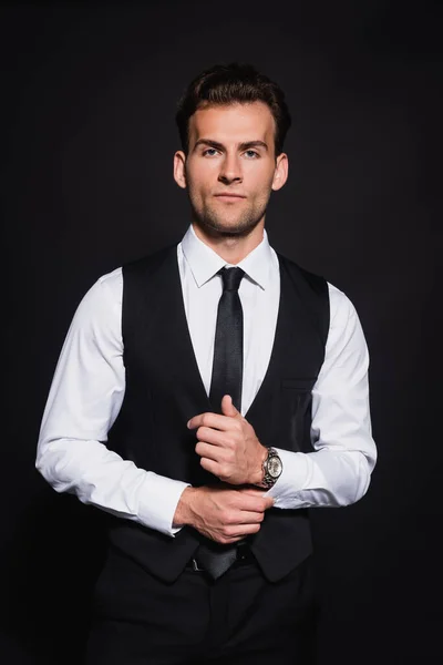 Serious and confident man buttoning sleeve of white shirt while looking at camera isolated on black — Stock Photo