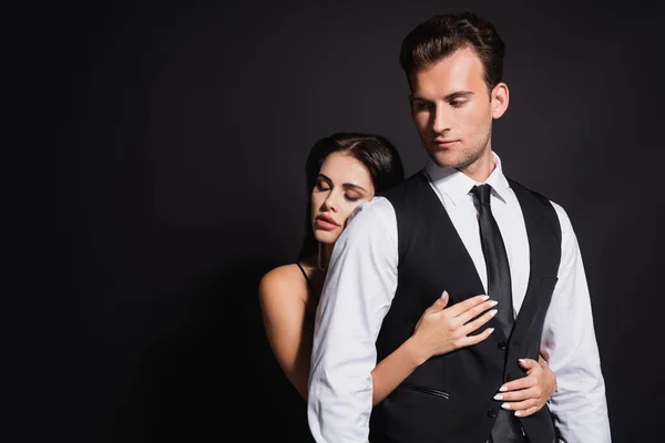 Joven morena mujer con los ojos cerrados abrazando elegante hombre aislado en negro - foto de stock