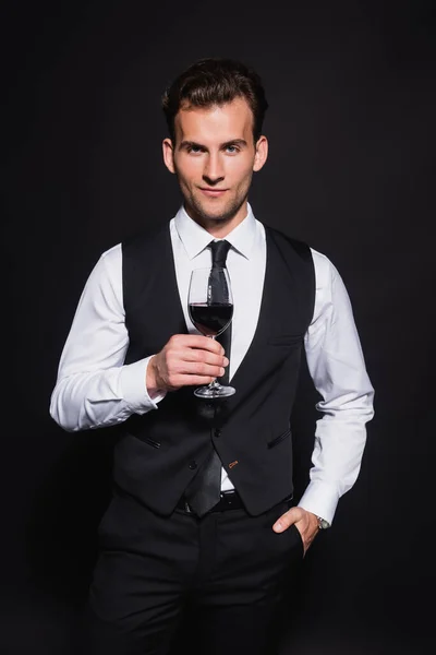 Vista frontale dell'uomo sorridente ed elegante che tiene in mano un bicchiere di vino rosso mentre è in piedi con la mano in tasca isolata sul nero — Foto stock