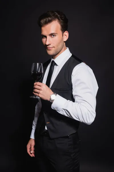 Hombre positivo y confiado mirando a la cámara mientras sostiene el vaso de vino tinto aislado en negro — Stock Photo