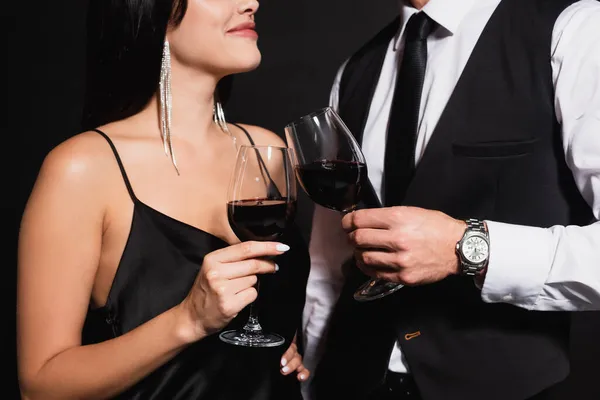 Recortado vista de sonriente mujer tintineo gafas con elegante hombre aislado en negro - foto de stock