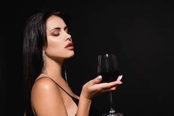 Sexy mujer con los ojos cerrados sosteniendo vaso de vino tinto aislado en negro - foto de stock