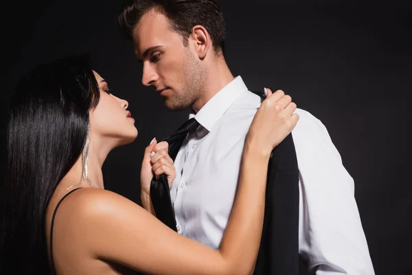 Passionate brunette woman pulling tie of man while undressing him isolated on black — Stock Photo