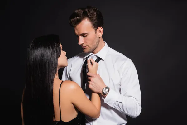 Hombre en camisa blanca mirando elegante morena mujer tocando su corbata aislada en negro - foto de stock