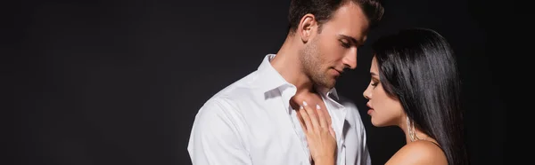 Bonita mujer morena cerca de hombre en camisa blanca desabotonada aislada en negro, pancarta - foto de stock