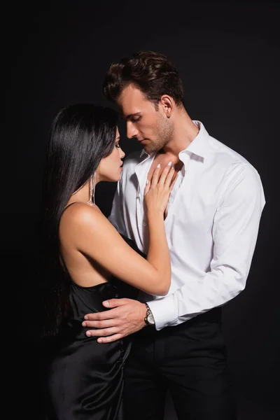 Hombre joven en camisa desabotonada abrazando a mujer elegante en vestido de seda aislado en negro - foto de stock