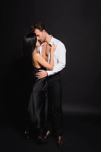 Vista completa de mujer morena en vestido elegante y hombre en camisa desabotonada abrazando en negro - foto de stock