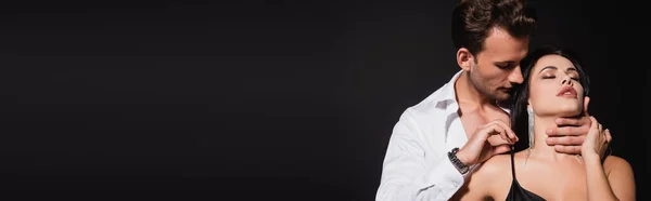 Hombre joven en camisa blanca desabotonada abrazando el cuello de la mujer sensual aislado en negro, bandera — Stock Photo